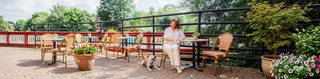 Woman sitting at a café by a canel with her dog tethered with a lead to her chair. 