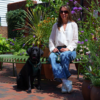 Black Labrador wearing a Forest Green organic cotton dog harness attached to matching Forest Green lead and waste bag holder, seated beside her owner in a planted urban courtyard